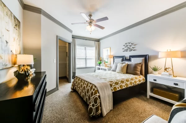 Bedroom with plush carpeting, a ceiling fan, large window, walk-in closet, and furnished with a king bed, nightstands, and a dresser